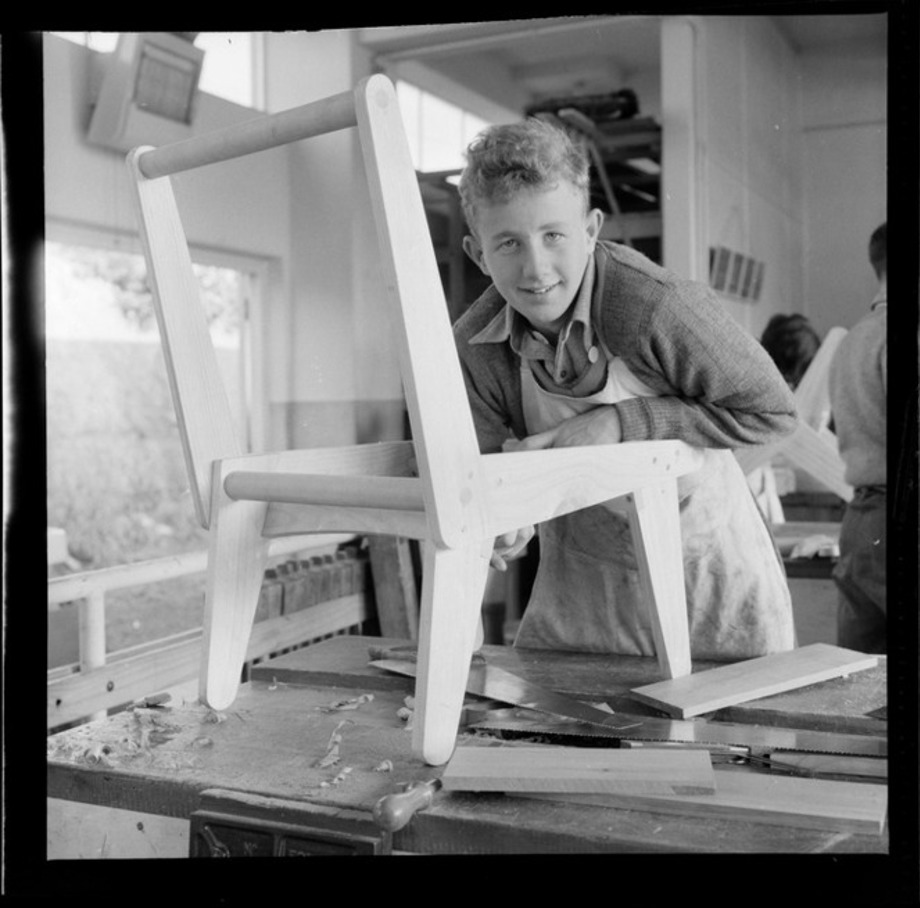 Wellington Technical College woodworking student, date unknown Young male student in apron leaning over wooden frame of a basic chair
